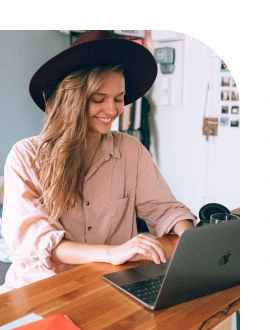 A smiling woman with long blond hair in a hat is working at a computer