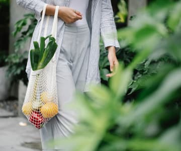 A woman in light-colored clothes walks with a bent hand, on which hangs a woven bag with vegetables.