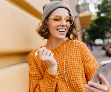 A smiling woman in sunglasses, a gray cap, and a knitted orange sweater stands on the street and, holding a smartphone in her hand, communicates via online communication.