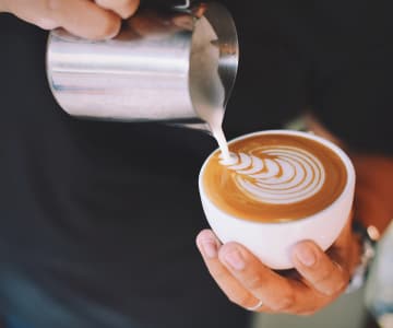 The barista holds a cup of coffee in his left hand and pours milky coffee with his right hand, creating a pattern.